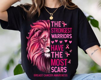Puede incluir: Una camiseta negra de manga corta con un gráfico de la cara de un león en el lado izquierdo y el texto "THE STRONGEST WARRIORS HAVE THE MOST SCARS" en rosa, acompañado de símbolos de lazos rosas y corazones pequeños, con "BREAST CANCER AWARENESS" debajo.