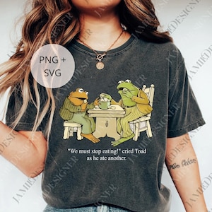 May include: Dark gray t-shirt featuring a whimsical illustration of two anthropomorphic frogs seated at a table. The frogs are dressed in clothing and the text reads "We must stop eating!" cried Toad as he ate another.