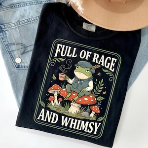 May include: Black t-shirt with a whimsical graphic featuring a frog in a cap and vest, holding a coffee cup, seated on a mushroom. The text reads "FULL OF RAGE AND WHIMSY" in a decorative frame. The shirt is laid on top of blue jeans.