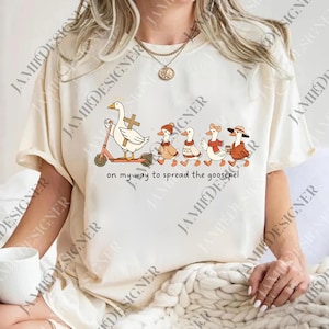 May include: Cream-colored t-shirt featuring a whimsical illustration of geese. One goose rides a pink scooter with a cross, leading a line of geese in various outfits. The text "on my way to spread the goosepel" is below the illustration.