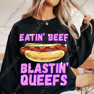 May include: Black sweatshirt with the text "EATIN' BEEF" above a hot dog illustration and "BLASTIN' QUEEFS" below. The text is in pink with a purple outline and white star accents. The hot dog is in a bun with ketchup.