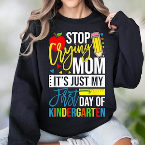 May include: A black long-sleeve sweatshirt featuring a graphic print with a red apple, a yellow pencil, and the text: STOP CRYING MOM IT'S JUST MY FIRST DAY OF KINDERGARTEN. The design includes colorful accents, a ruler graphic, and small hearts.
