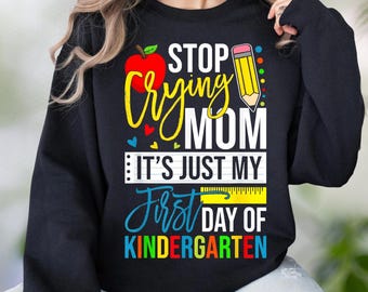 May include: A black long-sleeve sweatshirt featuring a graphic print with a red apple, a yellow pencil, and the text: STOP CRYING MOM IT'S JUST MY FIRST DAY OF KINDERGARTEN. The design includes colorful accents, a ruler graphic, and small hearts.