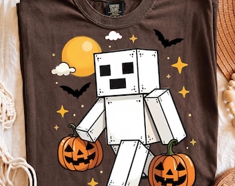 Puede incluir: Una camiseta marrón oscuro con un gráfico de un personaje blanco y cuadrado que parece un esqueleto, dos calabazas de Halloween naranjas, una luna llena amarilla, murciélagos negros y estrellas amarillas. Etiqueta Comfort Colors en el cuello.