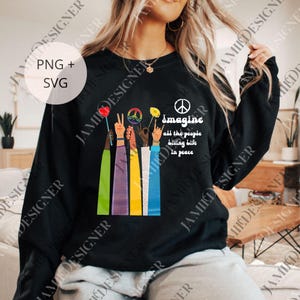 Puede incluir: Sudadera negra con un gráfico colorido de manos levantadas sosteniendo flores y símbolos de la paz. El texto "Imagine all the people living life in peace" está impreso en la parte delantera. El diseño incluye un símbolo de la paz.