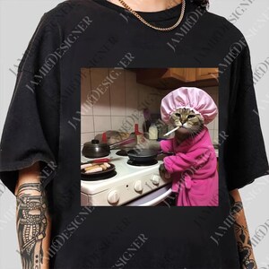 A person wearing a pink robe and a pink hair cap is cooking on a stove, with a cat wearing a pink hat sitting next to the stove.