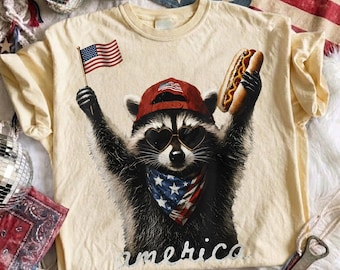 Puede incluir: Una camiseta color crema con el gráfico de un mapache que lleva una gorra roja, gafas de sol en forma de corazón y un pañuelo con estrellas y rayas. El mapache sostiene una pequeña bandera estadounidense y un perrito caliente. La palabra America aparece debajo.
