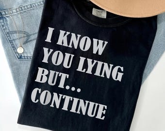 May include: A black short-sleeved t-shirt featuring the white printed text I KNOW YOU LYING BUT... CONTINUE. The shirt has a crew neckline and a Comfort Colors brand label visible on the inside collar area, presented with blue denim fabric.