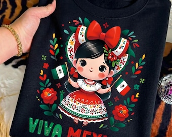 Puede incluir: Una sudadera negra con el gráfico de un personaje de dibujos animados de pelo negro con un gran lazo rojo, vistiendo un vestido blanco y rojo. El personaje sostiene dos pequeñas banderas mexicanas. El diseño incluye flores coloridas y el texto VIVA MEXICO.