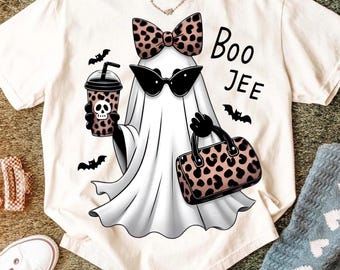 May include: A cream-colored t-shirt featuring a graphic of a white ghost wearing black cat-eye sunglasses and a leopard print bow. The ghost holds a leopard print cup with a skull and a leopard print handbag. The text reads Boo JEE with bats.