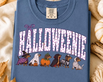 Puede incluir: Una camiseta azul de cuello redondo con el texto HALLOWEENIE en letras blancas con lunares morados. Debajo del texto hay seis perros salchicha con disfraces de Halloween, incluyendo una bruja, un fantasma, una calabaza, un vampiro, un esqueleto y una momia.