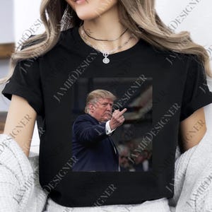 May include: Black t-shirt featuring a rectangular image of a man in a suit, raising his middle finger. The image is centered on the shirt. The shirt has short sleeves and a crew neck. The text "JAMII DESIGNER" is printed on the shirt.