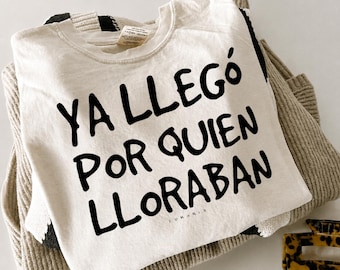 Puede incluir: Una camiseta de cuello redondo color crema con el texto negro "YA LLEGÓ POR QUIEN LLORABAN" impreso en el frente. La camiseta está sobre un suéter de punto a rayas beige y negro, con una pequeña pinza para el cabello de carey cerca.