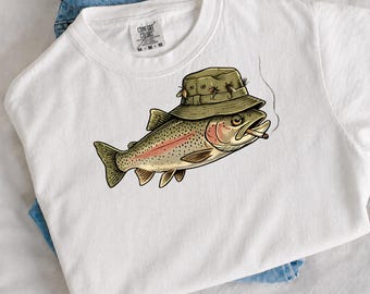 May include: A white t-shirt featuring a graphic of a trout wearing an olive green fishing hat with small flies attached to the brim. The fish has a cigarette in its mouth, with a thin line of smoke rising upward. The shirt has a Comfort Colors label.