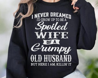 Puede incluir: Una sudadera negra de manga larga con cuello redondo que presenta el texto en blanco: I NEVER DREAMED I'D GROW UP TO BE A Spoiled WIFE OF A Grumpy OLD HUSBAND BUT HERE I AM, KILLIN' IT. La prenda tiene un corte estándar con puños y cuello acanalados.