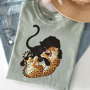Könnte beinhalten: Salbeigrünes T-Shirt mit einem Aufdruck einer schwarzen Panther und eines Leoparden. Der Leopard hat orange und schwarze Flecken. Das T-Shirt wird mit einer Jeans, einer Halskette und einem beigen Hut kombiniert. Das T-Shirt hat den Text "Comfort Colors".