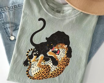 May include: Sage green t-shirt featuring a graphic of a black panther embracing a leopard. The leopard has orange and black spots, with its mouth open. The shirt is paired with blue jeans, a necklace, and a tan hat.