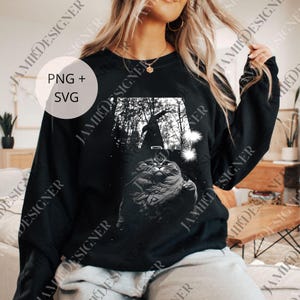May include: Black sweatshirt featuring a grayscale graphic of a cat wearing a witch's hat, set against a forest backdrop. The design includes the text "PNG + SVG" in a white circle. A cozy, long-sleeved top perfect for Halloween or cat lovers.