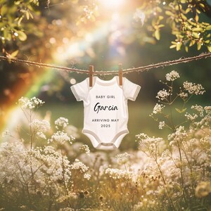 May include: A white baby onesie hanging on a clothesline in a field of white flowers. The onesie has the text "BABY GIRL Garcia ARRIVING MAY 2025" printed on it.