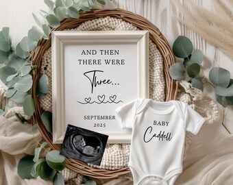 May include: A white framed sign with the text "And then there were Three... September 2025" with three small hearts. A white baby onesie with the text "Baby Caddell" is next to the sign. An ultrasound image is in front of the sign.