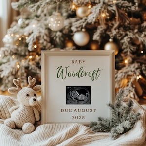May include: A white frame with a brown border, announcing the arrival of "Baby Woodcroft" with a sonogram image and the due date "Due August 2025". The frame is placed on a white blanket with a brown knitted reindeer toy in the foreground.