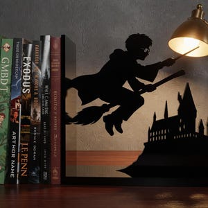 Könnte beinhalten: Schwarze Metall-Buchstützen mit einer Silhouette einer Figur auf einem Besen, die auf ein Schloss zufliegt. Bücher sind zwischen den Buchstützen platziert. Rechts von den Buchstützen befindet sich eine goldfarbene Schreibtischlampe. Die Bücher tragen Titel wie "The Hobbit" und "Exodus".