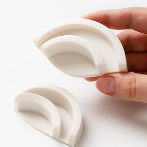 Puede incluir: Dos objetos semicirculares de color blanquecino en capas. Uno es sostenido por una mano, el otro descansa debajo. Los objetos tienen un acabado liso y mate y un dise&ntilde;o minimalista. Parecen ser objetos peque&ntilde;os, posiblemente decorativos o funcionales.