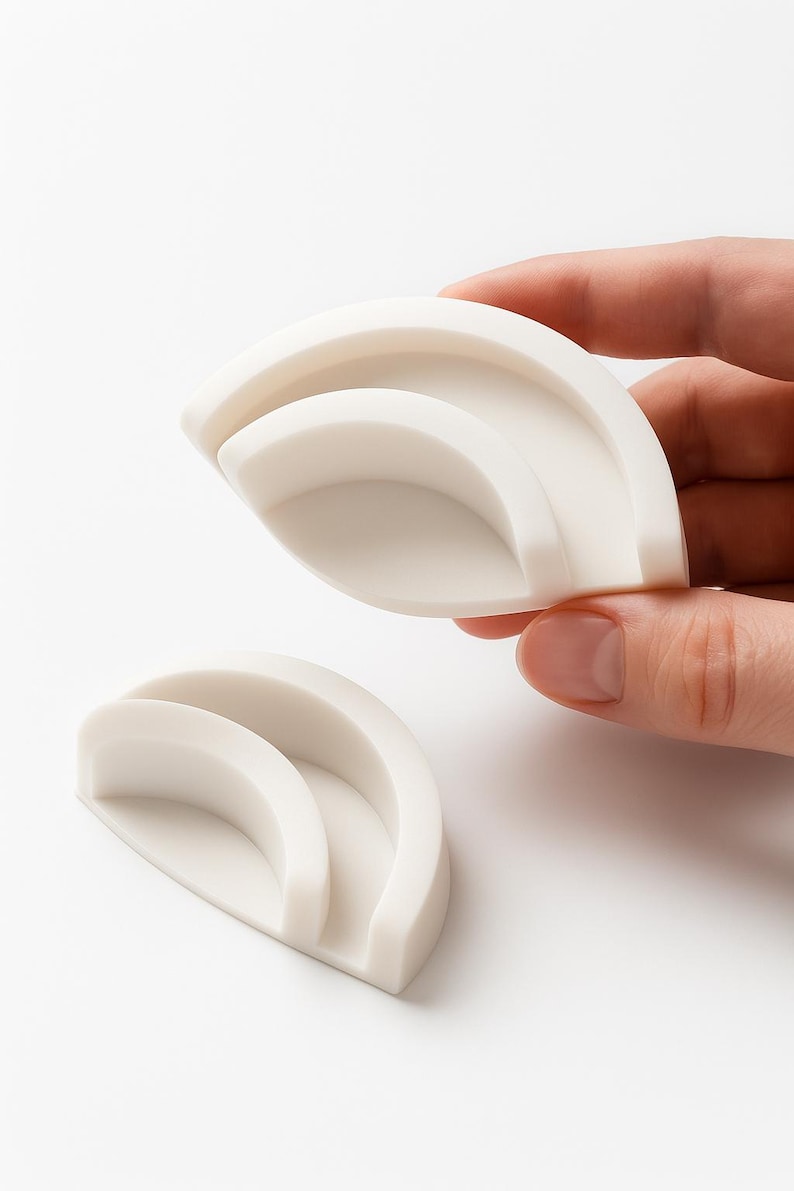 Puede incluir: Dos objetos semicirculares de color blanquecino en capas. Uno es sostenido por una mano, el otro descansa debajo. Los objetos tienen un acabado liso y mate y un dise&ntilde;o minimalista. Parecen ser objetos peque&ntilde;os, posiblemente decorativos o funcionales.