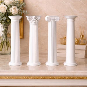 Può includere: Quattro colonne decorative bianche con diversi disegni di capitelli. Sono disposte su una superficie di marmo, con uno specchio con cornice dorata e un vaso di rose bianche sullo sfondo. Queste colonne sono un elemento architettonico classico.