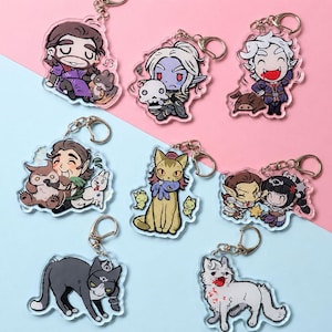 Puede incluir: Una colección de ocho llaveros de acrílico transparente con coloridas ilustraciones de dibujos animados de varios personajes de fantasía, incluyendo humanoides, un gato y un lobo, cada uno unido a una anilla de metal plateado.