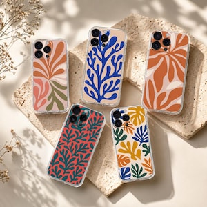 May include: Five clear phone cases with colorful abstract floral designs. The cases feature various patterns in shades of orange, pink, green, blue, and yellow. The cases are displayed on a textured, beige surface, with a sprig of greenery in the corner.