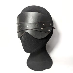 Puede incluir: Una venda de cuero negro con remaches plateados. La venda está sobre una cabeza de maniquí negra. El diseño presenta una forma curva para adaptarse a la cara y una correa para sujetarla.