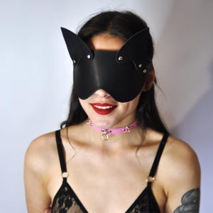 May include: Black leather cat-ear mask with silver rivets. A pink studded collar with a silver ring and a black lace bra with adjustable straps. The person is smiling with red lipstick.