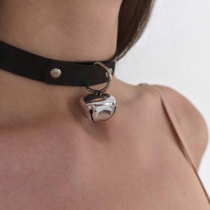 Könnte beinhalten: Schwarze Leder-Halskette mit einem silbernen Glockenanhänger. Die Halskette wird mit einem silbernen Druckknopf verschlossen. Die Glocke ist glänzend und reflektierend.