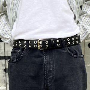 May include: Black leather belt with silver grommets and a silver buckle. The belt is worn with black denim jeans and a white t-shirt. The belt has two rows of grommets and a rectangular silver buckle.