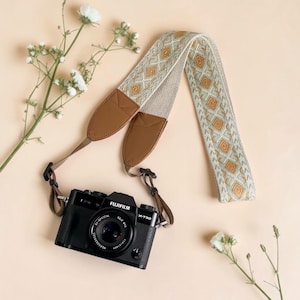 Puede incluir: Una cámara Fujifilm X-T30 negra con una correa estampada. La correa presenta un diseño geométrico beige y dorado con extremos de cuero marrón. La cámara y la correa están sobre una superficie de color melocotón claro, con flores blancas.