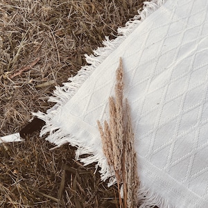 May include: A white, textured blanket with a diamond pattern and fringed edges, resting on a bed of dry grass. A bundle of dried, beige-colored grass is placed on top of the blanket, creating a rustic, natural aesthetic.