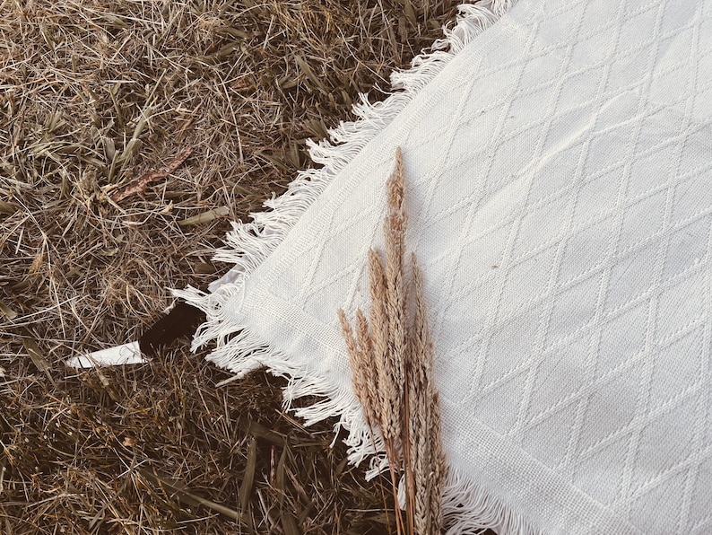 May include: A white, textured blanket with a diamond pattern and fringed edges, resting on a bed of dry grass. A bundle of dried, beige-colored grass is placed on top of the blanket, creating a rustic, natural aesthetic.