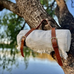 Könnte beinhalten: Eine aufgerollte weiße Picknickdecke mit Fransen, befestigt mit zwei braunen Lederriemen mit Messingschnallen. Die Decke hängt an einem Ast, mit einem See und blauem Himmel im Hintergrund.