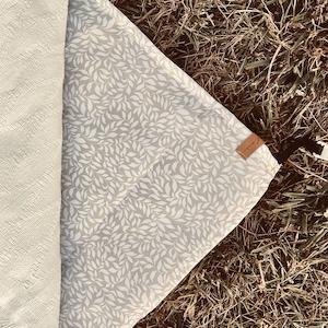 May include: A rolled-up cream-colored blanket and a gray picnic blanket with a white leaf pattern are displayed on grass. A small brown tag is attached to the picnic blanket. The blankets are ideal for outdoor use.