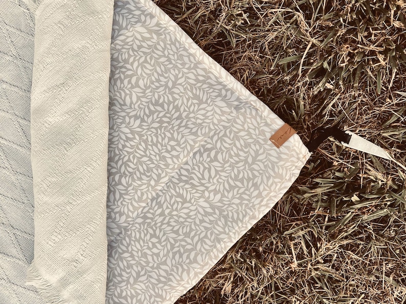 May include: A rolled-up cream-colored blanket and a gray picnic blanket with a white leaf pattern are displayed on grass. A small brown tag is attached to the picnic blanket. The blankets are ideal for outdoor use.