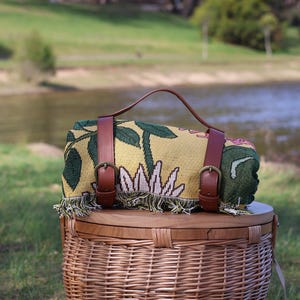 May include: Rolled picnic blanket with a yellow floral pattern, brown leather straps, and a handle. The blanket is resting on a wicker picnic basket with a wooden lid. The scene is set outdoors near a body of water.