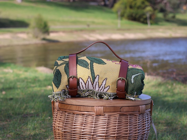 May include: Rolled picnic blanket with a yellow floral pattern, brown leather straps, and a handle. The blanket is resting on a wicker picnic basket with a wooden lid. The scene is set outdoors near a body of water.