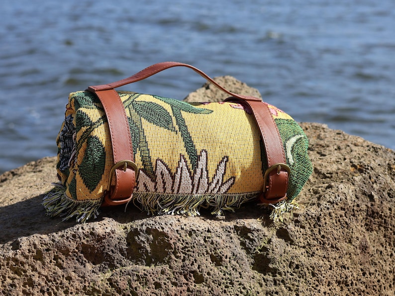 May include: Rolled-up picnic blanket with a floral pattern in yellow, green, and white. It features brown leather straps and a handle for easy carrying. The blanket is fringed at the edges and rests on a rock near the water.