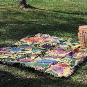 May include: A yellow floral picnic blanket with a fringed edge is spread on green grass. The blanket features a colorful pattern of flowers in shades of pink, blue, orange, and green. A wicker picnic basket sits nearby.