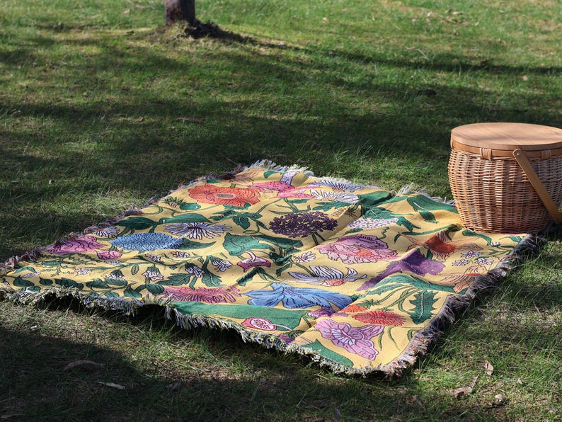 May include: A yellow floral picnic blanket with a fringed edge is spread on green grass. The blanket features a colorful pattern of flowers in shades of pink, blue, orange, and green. A wicker picnic basket sits nearby.