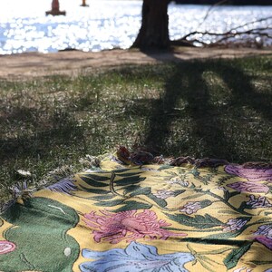 May include: A yellow floral patterned blanket with pink, blue, and green accents is spread on the grass. The blanket is fringed and has a botanical design. The background shows a lake and trees.