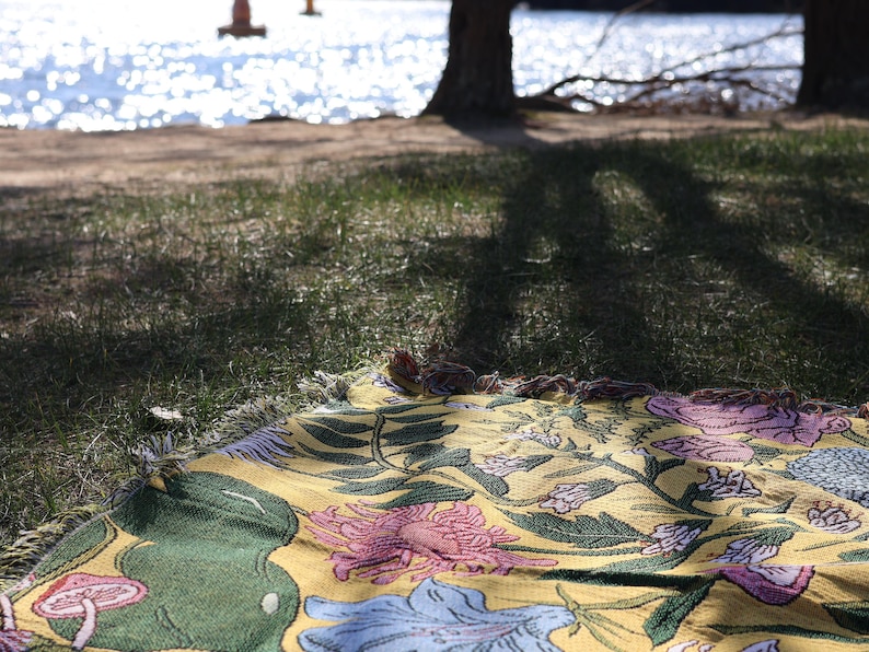 May include: A yellow floral patterned blanket with pink, blue, and green accents is spread on the grass. The blanket is fringed and has a botanical design. The background shows a lake and trees.