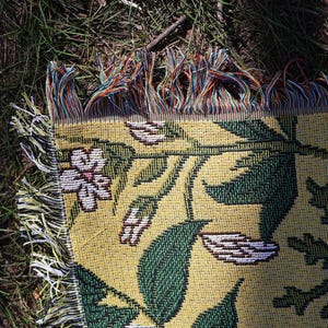 May include: A woven throw blanket with a floral pattern. The blanket features a yellow background with green leaves and white flowers. The edges have colorful fringe.