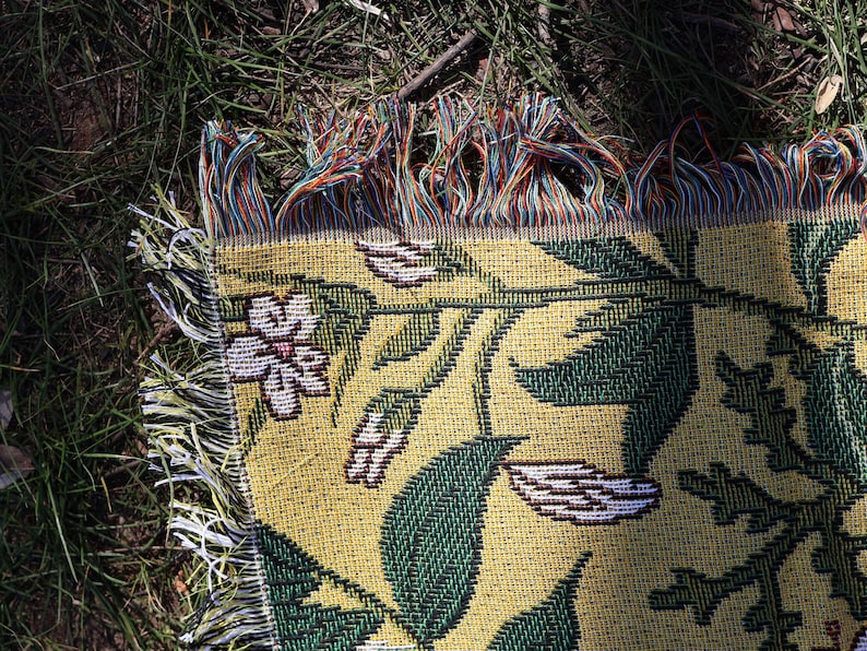 May include: A woven throw blanket with a floral pattern. The blanket features a yellow background with green leaves and white flowers. The edges have colorful fringe.